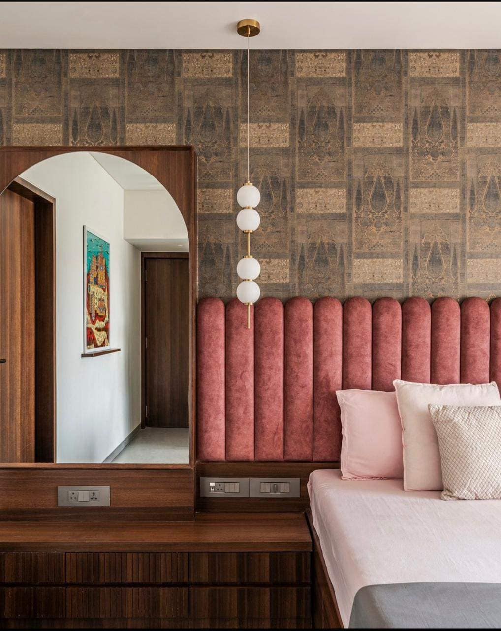 Elegant bedroom interior design by Olangana Design in Bangalore featuring a pink upholstered headboard, a stylish wooden dresser with an arched mirror, modern pendant lighting, and patterned wallpaper, showcasing a sophisticated residential space.