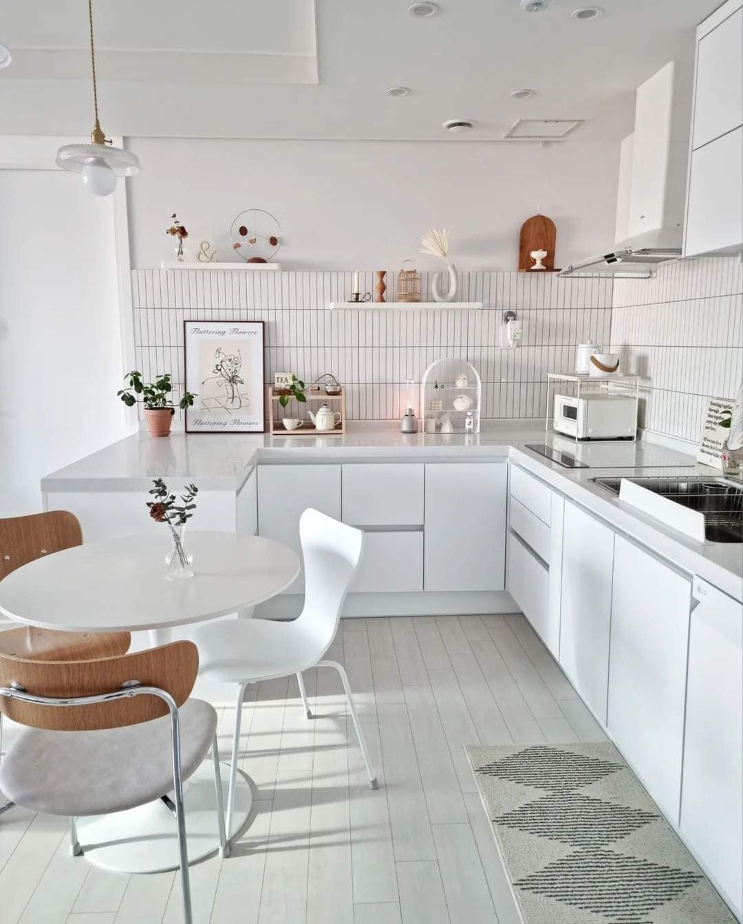 Modern minimalist all-white kitchen and dining area interior design by Olangana in Bangalore, featuring sleek white cabinetry, a U-shaped layout, and a round dining table with mixed chairs.
