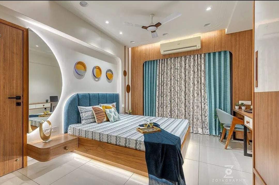 Modern bedroom interior design by Olangana Design in Bangalore, featuring a blue upholstered headboard, wooden bed, illuminated mirror accent wall, and teal curtains for a luxurious feel.