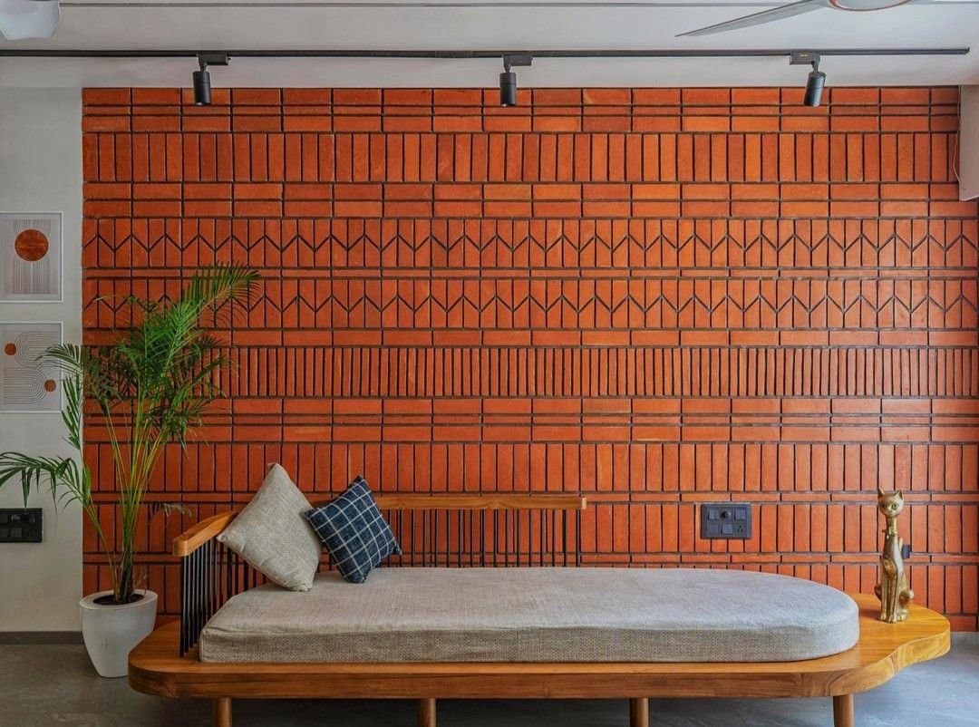 Modern living room interior design by Olangana in Bangalore, showcasing a striking terracotta brick accent wall, a sleek wooden daybed, and elegant track lighting.