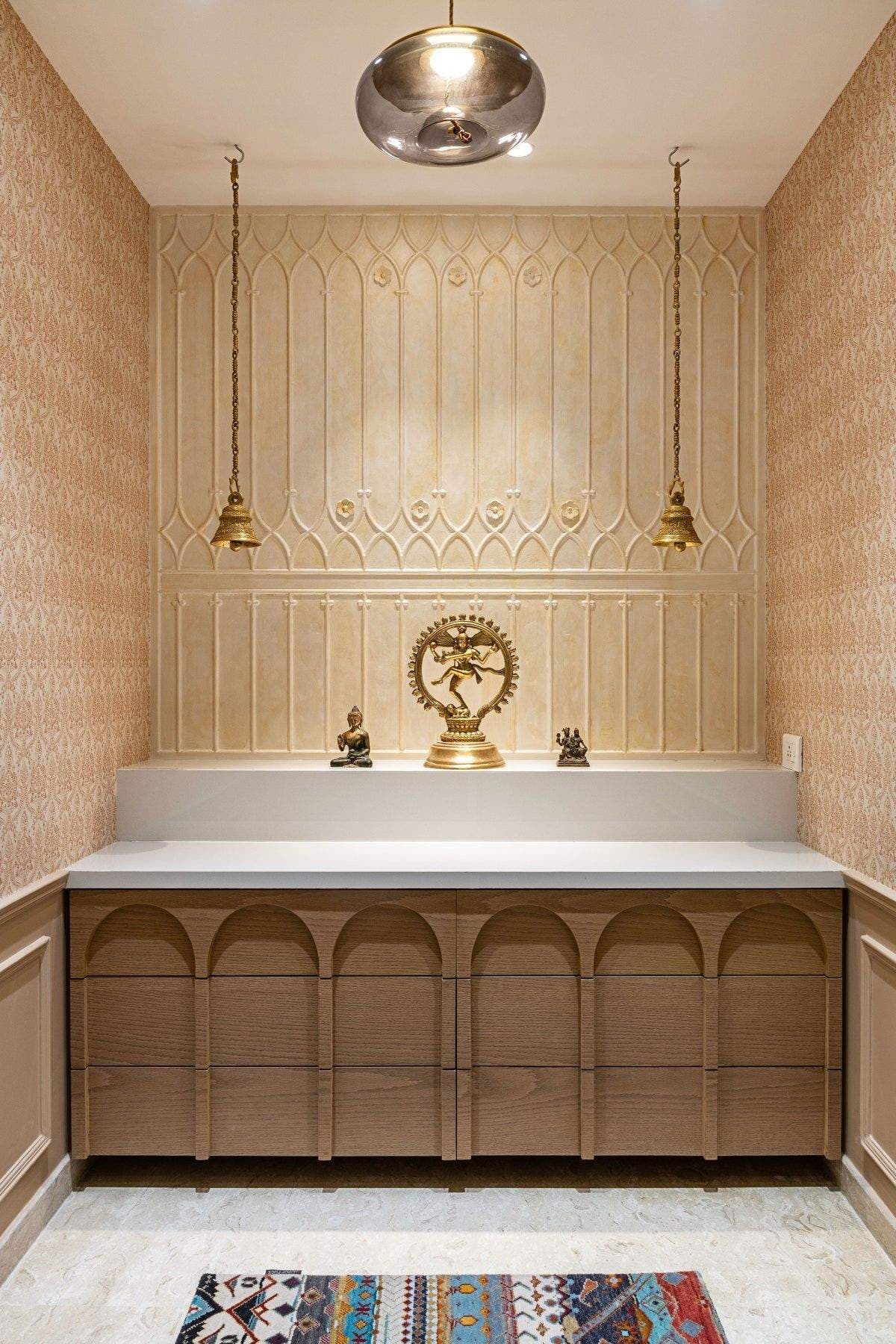 Elegant puja room interior design by Olangana in Bangalore, featuring warm neutral tones, intricately carved wall paneling, brass hanging bells, and modern light wood cabinetry with arched details, adorned with deity statues.