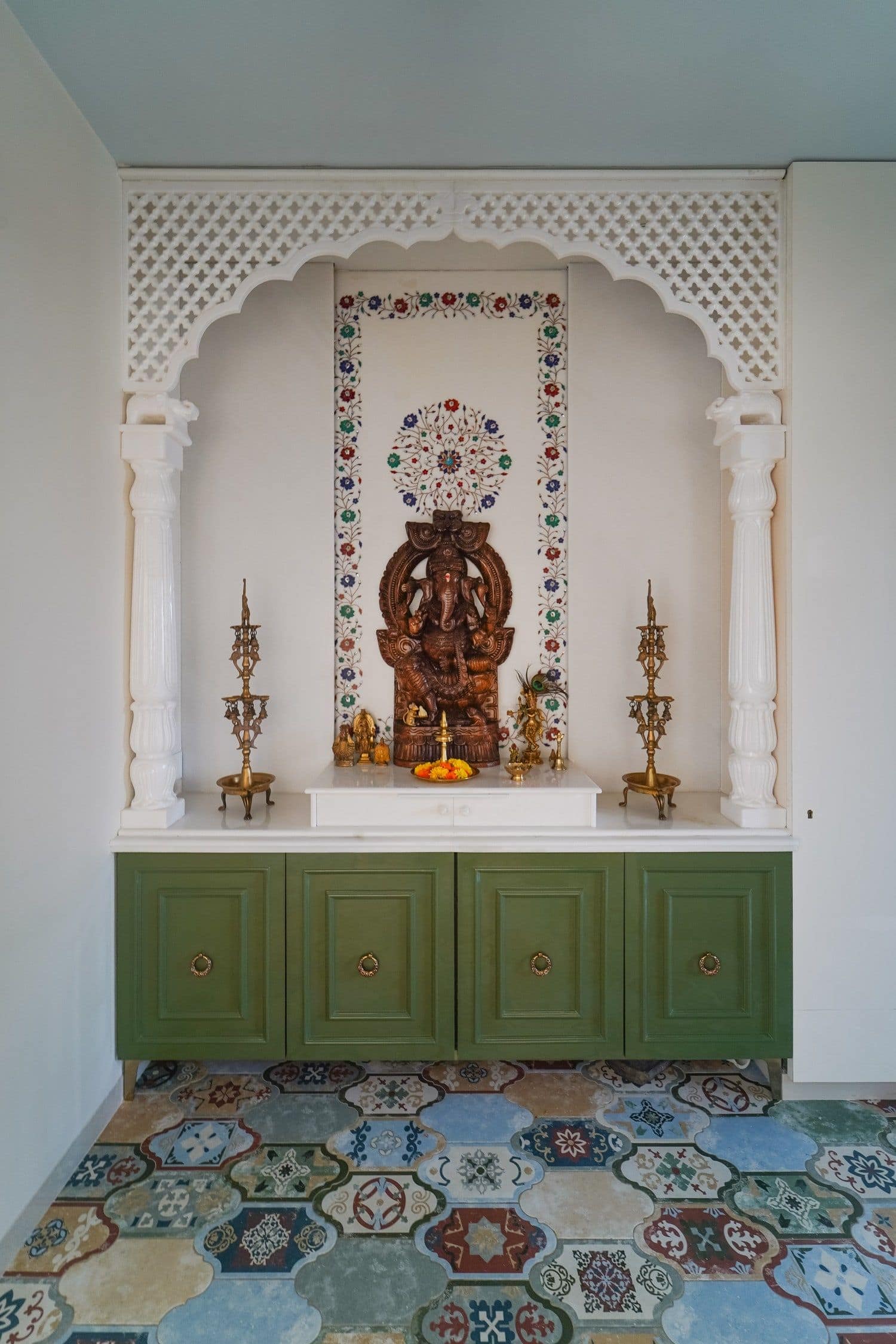 Elegant home temple interior design by Olangana in Bangalore, featuring a white marble arch, intricate inlay work, a Ganesha idol, green storage cabinets, and patterned floor tiles.