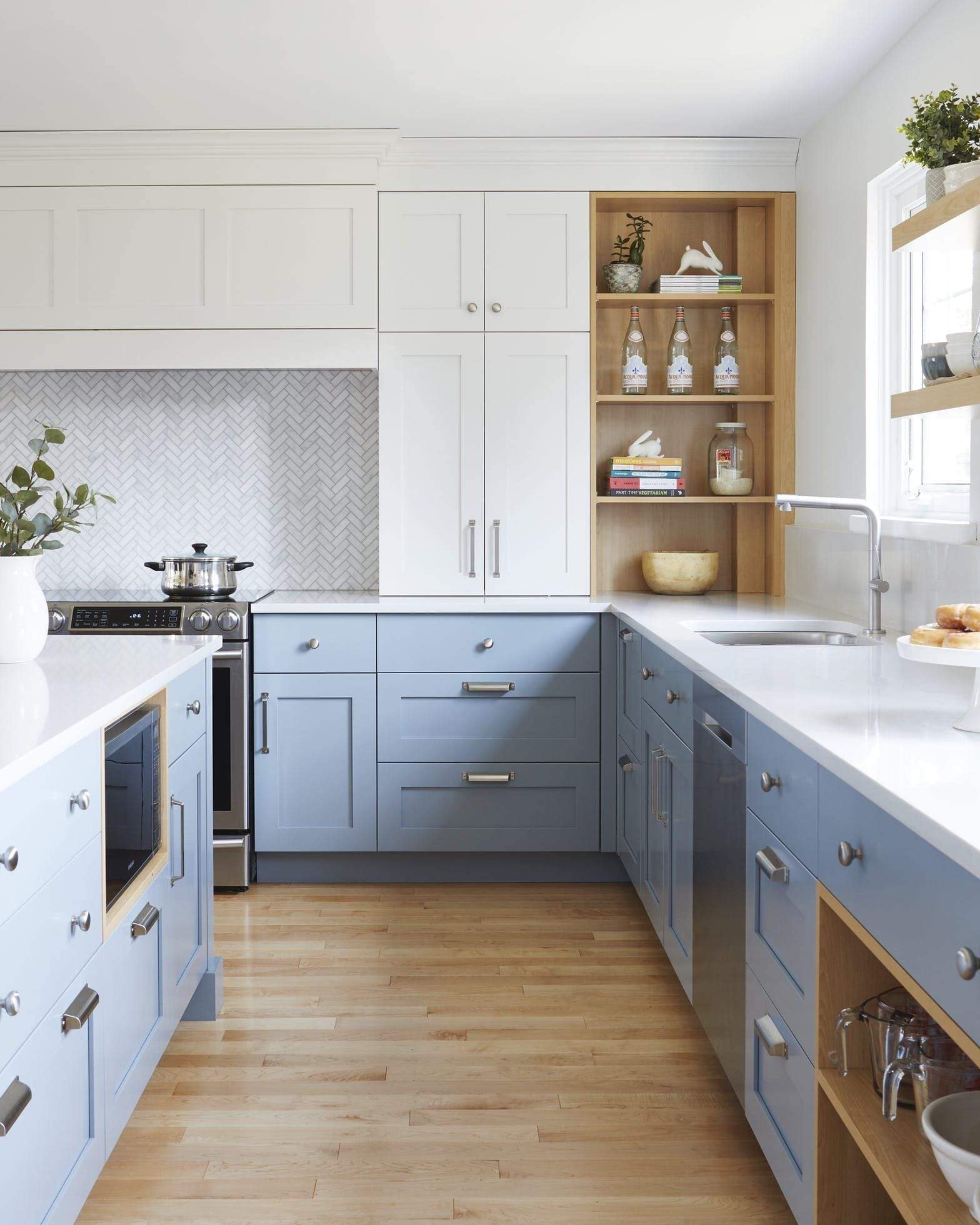 Modern kitchen interior design by Olangana in Bangalore, featuring light blue and white cabinetry, white quartz countertops, a herringbone tile backsplash, and integrated wooden shelving.