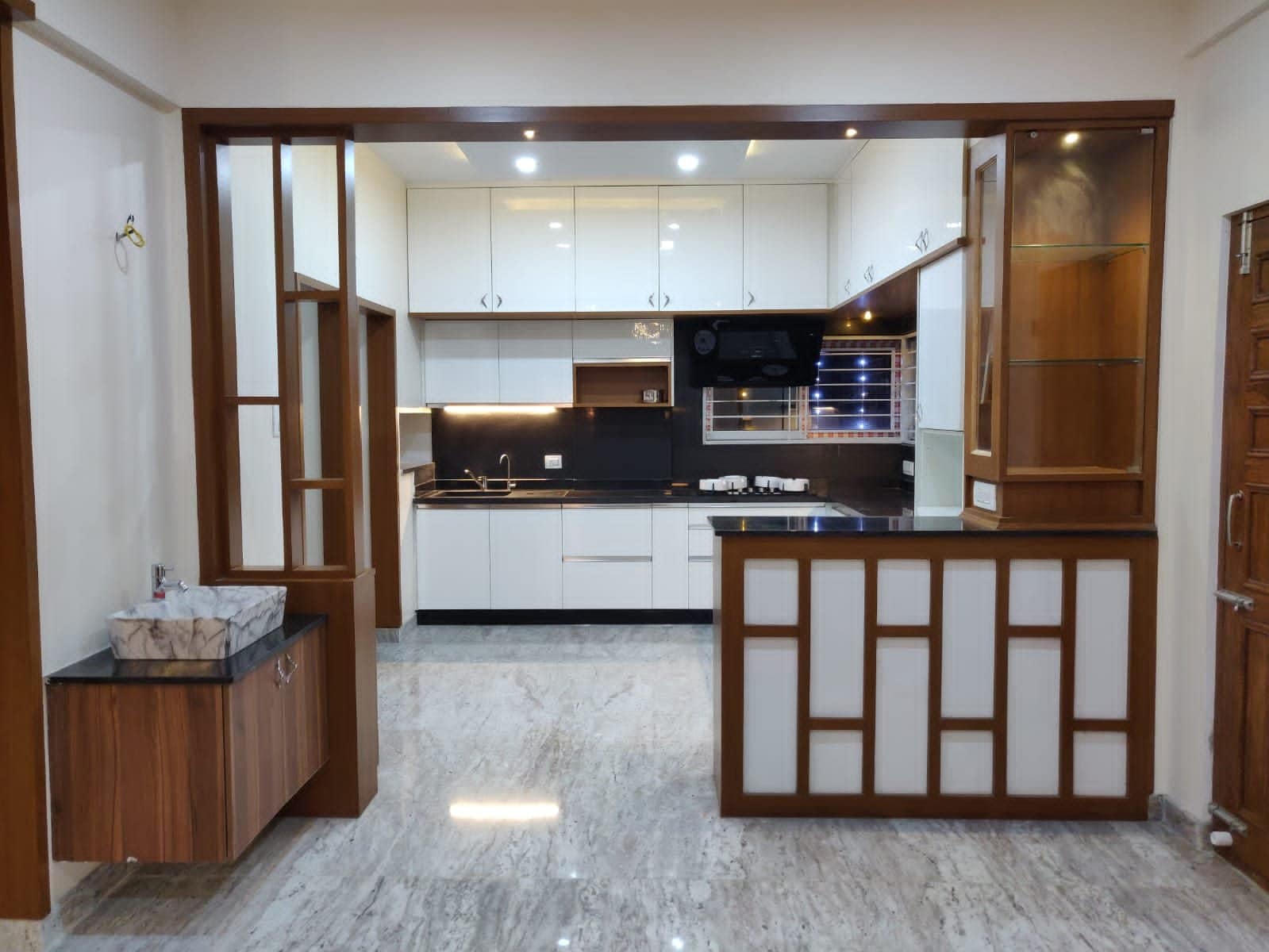 Modern kitchen interior design by Olangana in Bangalore, featuring sleek cream and taupe handleless cabinets, a light speckled countertop, and a black built-in hob with a slanted chimney.