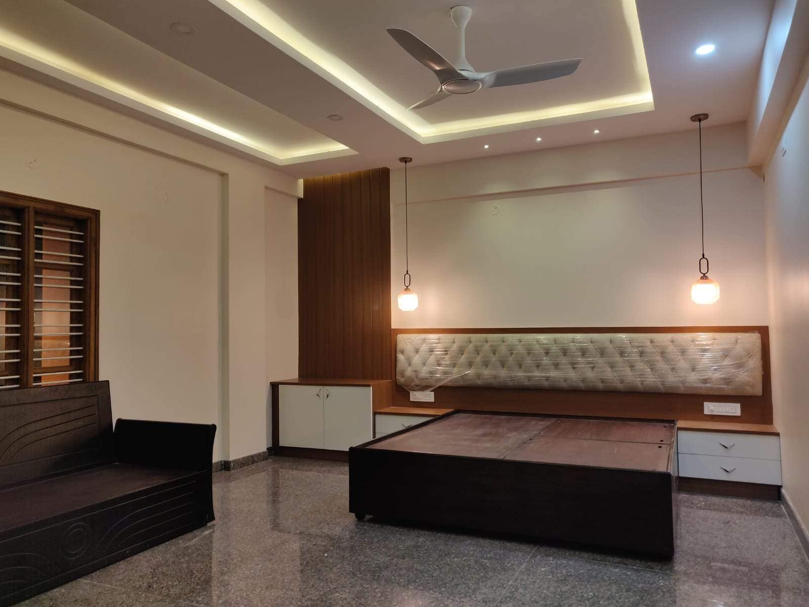 A modern bedroom interior in Bangalore by Olangana Design in its handover phase, featuring a dark wood bed frame, a plastic-wrapped upholstered headboard, contemporary pendant lights, false ceiling with cove lighting, and a sofa, awaiting final styling by Olangana Design.