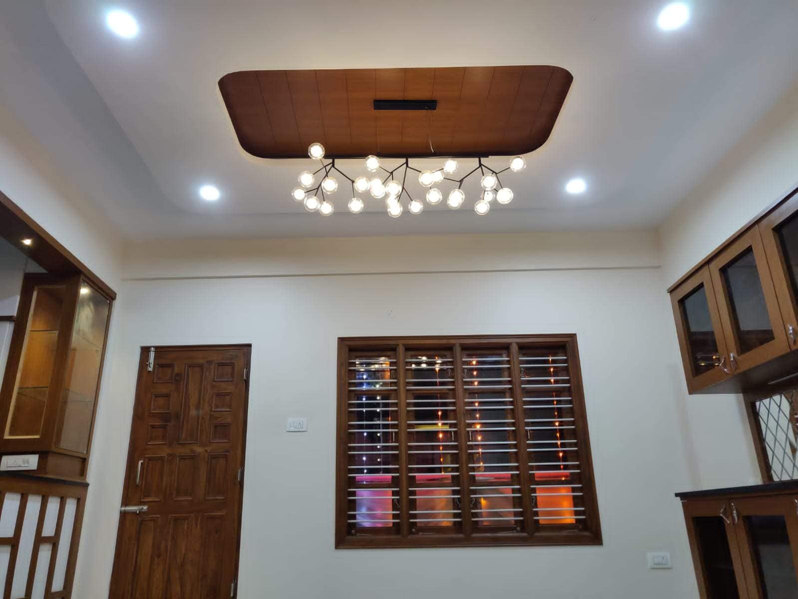 Modern living room interior design by Olangana in Bangalore, showcasing a custom false ceiling with a unique branch-like chandelier, elegant wooden window, and integrated wooden cabinetry.
