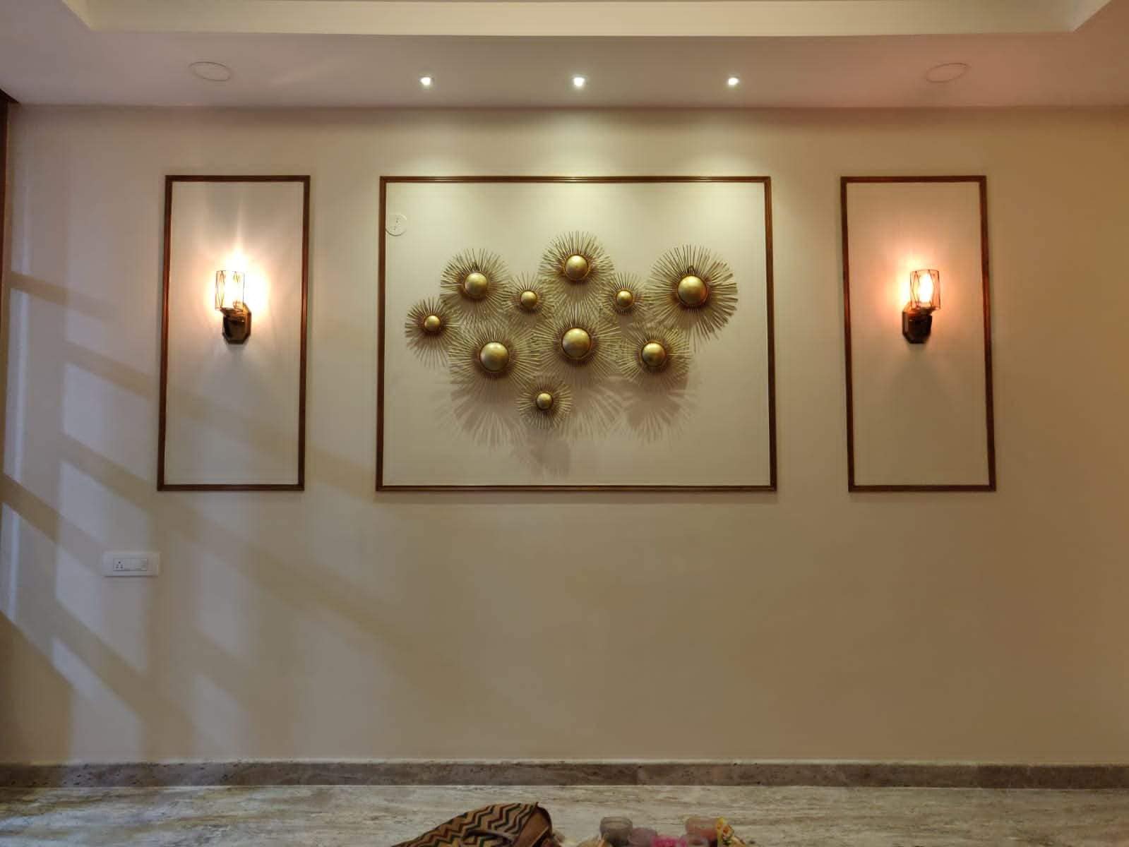 Elegant wall decor interior design by Olangana in Bangalore, featuring a striking gold sunburst metal art piece flanked by two illuminated wall sconces within dark frames, creating a sophisticated ambiance.