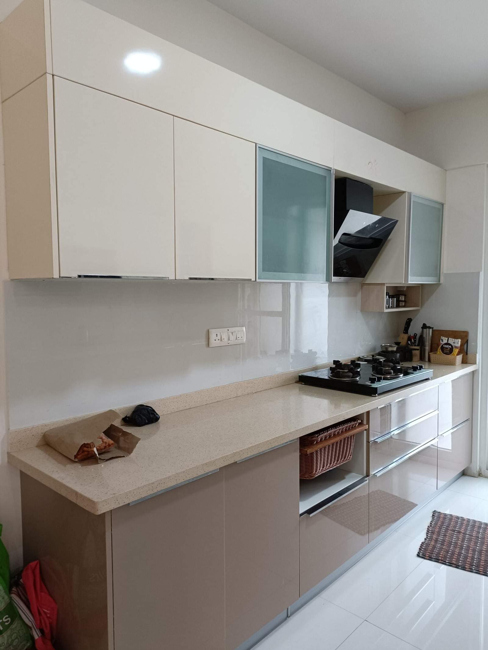 Modern kitchen interior design by Olangana in Bangalore, featuring sleek cream and taupe cabinetry, a speckled countertop, integrated hob and chimney, and stylish glass-fronted storage.
