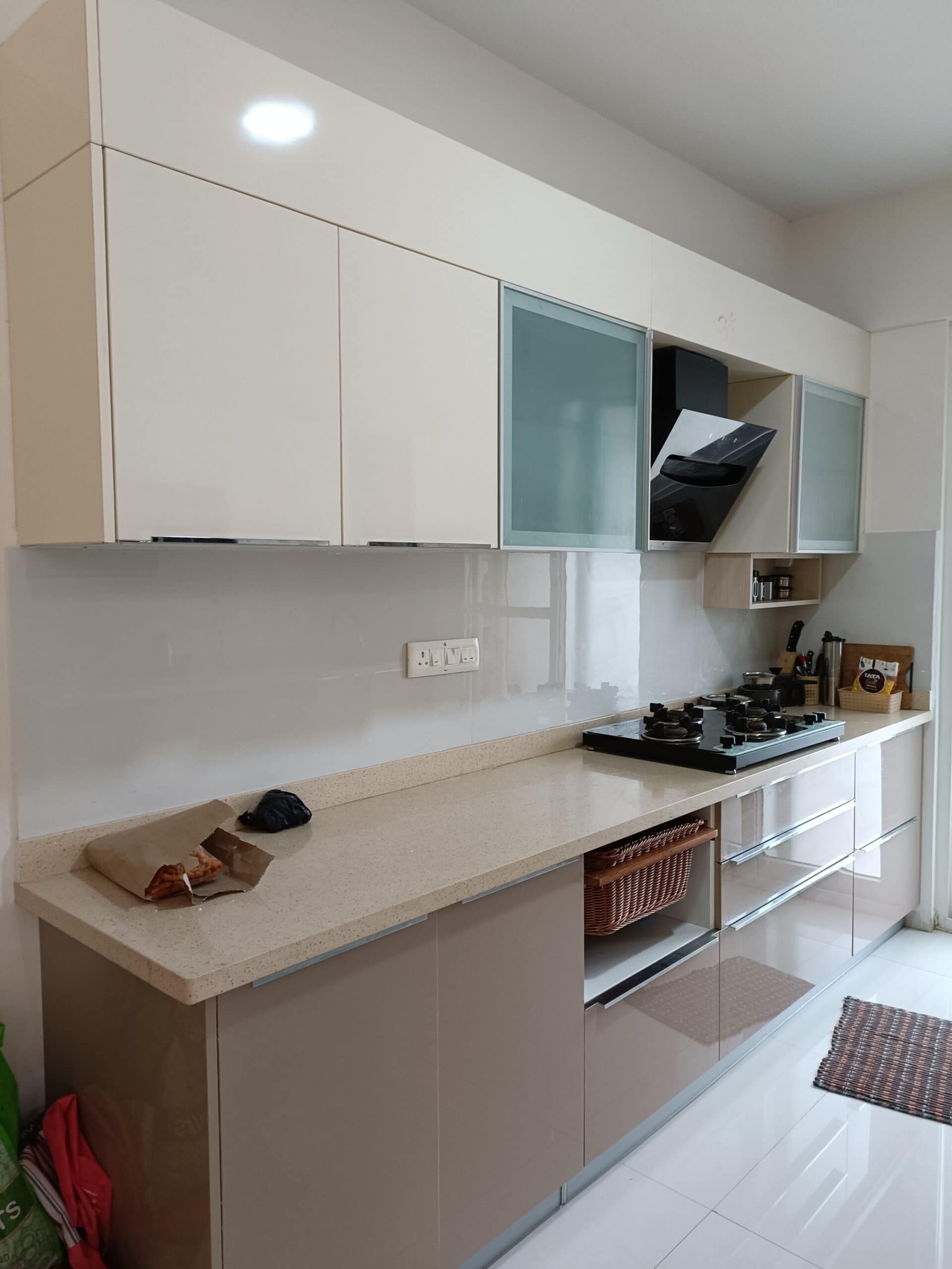 Modern kitchen interior design by Olangana in Bangalore, featuring sleek cream and taupe handleless cabinets, a light speckled countertop, and a black built-in hob with a slanted chimney.
