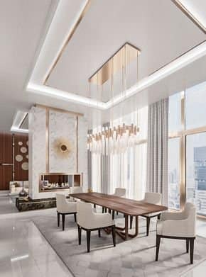 Luxury modern dining room interior design by Olangana Design in Bangalore, featuring a long wooden dining table, upholstered chairs, a striking gold chandelier, and a marble fireplace with a golden sunburst accent. Large windows offer a city view.