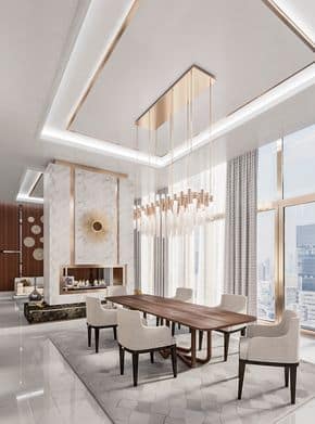 Luxury modern dining room interior design by Olangana Design in Bangalore, featuring a long wooden dining table, upholstered chairs, a striking gold chandelier, and a marble fireplace with a golden sunburst accent. Large windows offer a city view.
