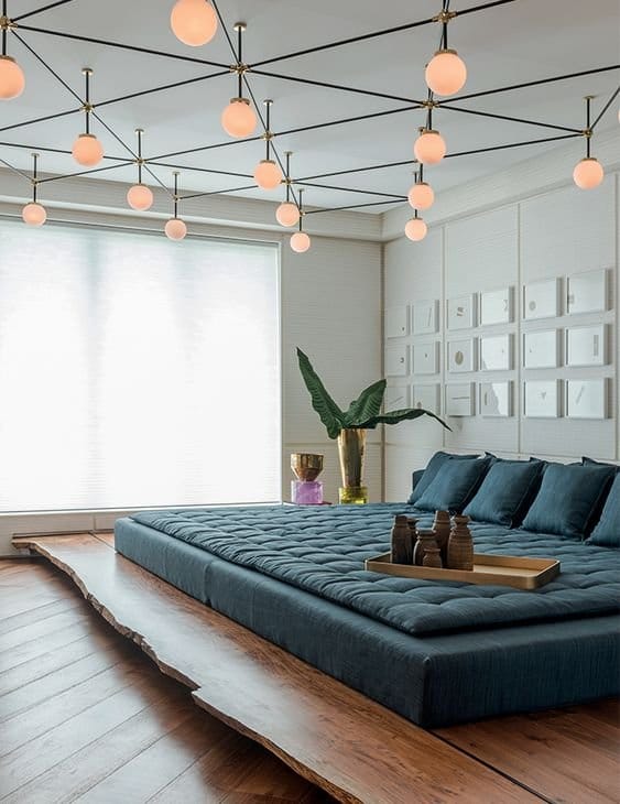 Luxurious bedroom interior design by Olangana in Bangalore, featuring a dark blue low-profile bed on a live-edge wooden platform, geometric ceiling lighting, and textured wall art.