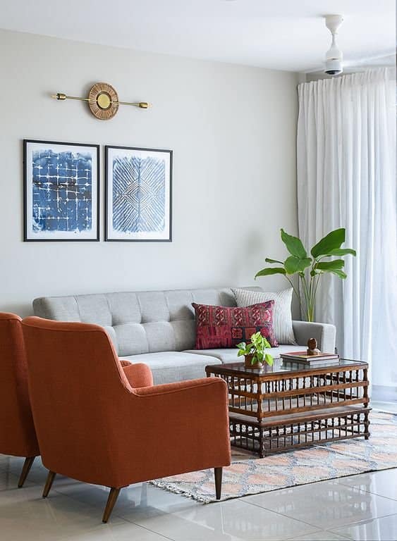 Elegant living room interior design by Olangana in Bangalore, featuring a grey tufted sofa, burnt orange armchairs, striking blue abstract art, and a classic wooden coffee table.