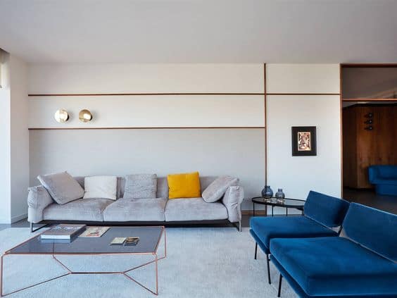 Modern living room interior design by Olangana in Bangalore, featuring a grey velvet sofa, vibrant blue armchairs, a geometric copper coffee table, and elegant wall paneling with gold sconces.