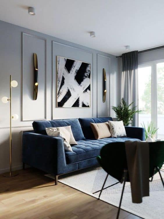 Elegant living room interior design by Olangana in Bangalore, featuring a luxurious navy blue velvet sofa, striking black and white abstract art, and sophisticated gold lighting against light grey paneled walls.