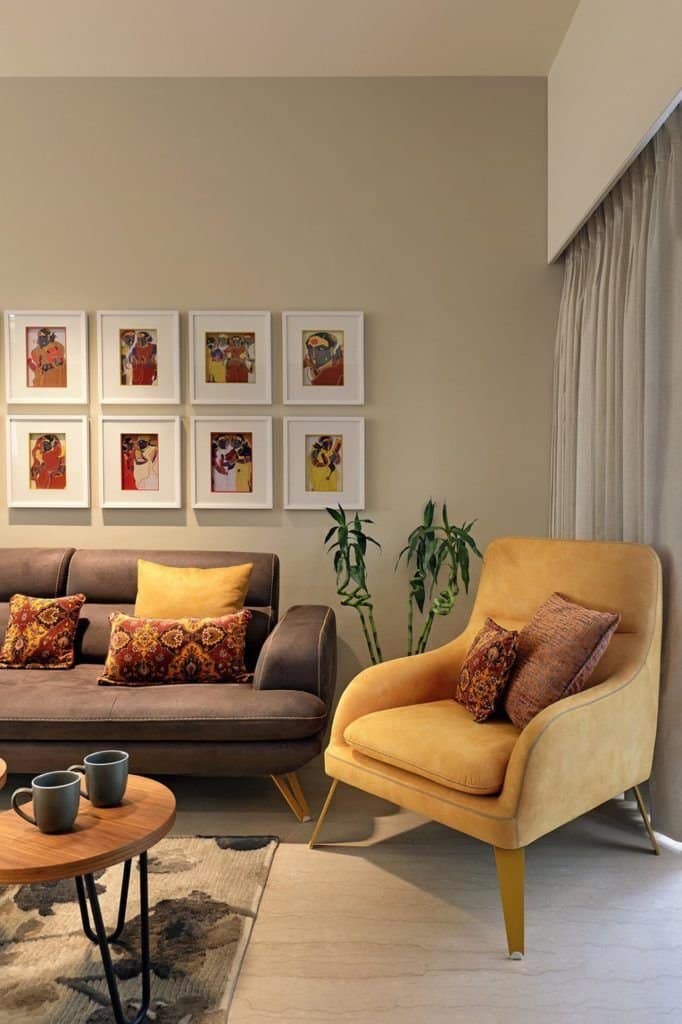 Warm and inviting living room interior design by Olangana in Bangalore, showcasing a brown sofa, a mustard yellow armchair, a gallery wall of vibrant artworks, and a round wooden coffee table, creating a cozy and elegant space.