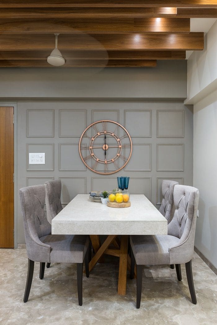 Modern dining interior design by Olangana Design in Bangalore featuring a grey paneled wall, stylish dining table, tufted chairs, and a unique rose gold clock.