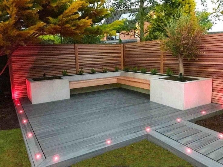 Modern outdoor patio interior design by Olangana in Bangalore, featuring grey composite decking, built-in wooden bench seating, concrete planters, and ambient red LED lighting for a sophisticated ambiance.