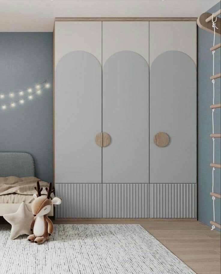 Elegant children's bedroom interior design by Olangana in Bangalore, showcasing a bespoke light grey and white wardrobe with arched details and fluted lower panels, alongside a cozy bed and playful decor.