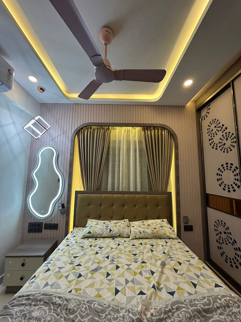 Modern master bedroom with warm lighting, sleek sliding wardrobe, POP ceiling with fan, stylish bed setup, curtains, and dressing table with mirror lighting by Olangana Design’s.