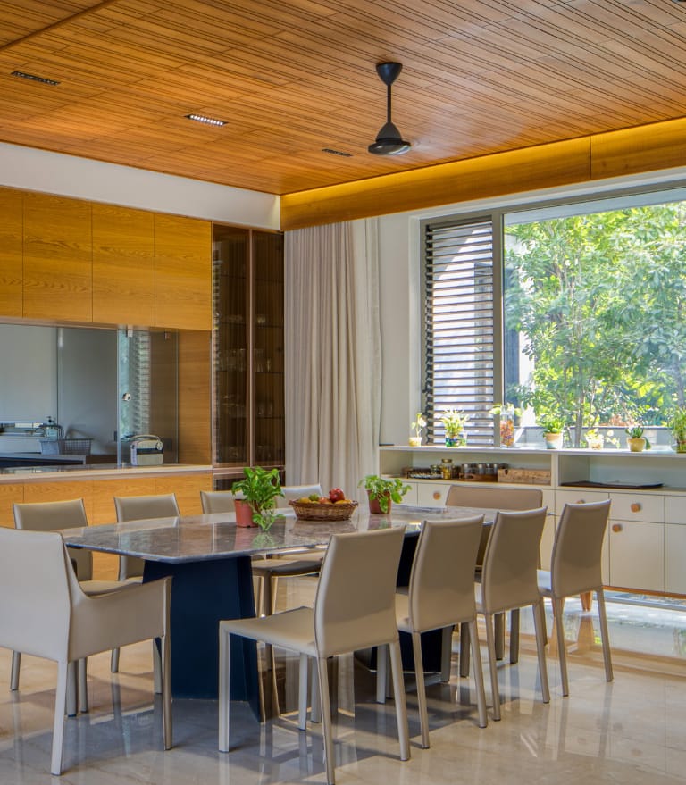 Modern dining room interior by Olangana Design in Bangalore featuring a sleek marble dining table, elegant beige chairs, and a warm wooden slatted ceiling with ample natural light.