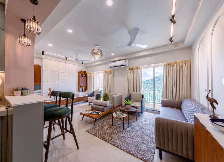 Elegant open-concept living room interior design by Olangana in Bangalore, featuring a modern bar counter with green velvet stools, comfortable neutral seating, and scenic window views.