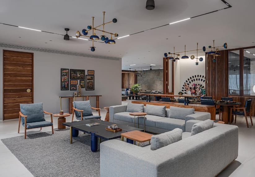 Modern open-plan lounge interior design by Olangana in Bangalore, featuring light grey sectional sofas, blue accent armchairs, a dark coffee table, and unique gold and blue chandeliers.