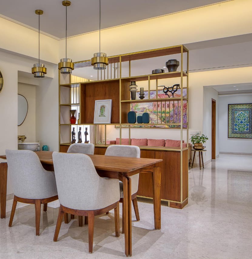 Elegant dining interior design by Olangana Design in Bangalore featuring a wooden dining table, light grey upholstered chairs, and a stylish brass and wood shelving unit with modern decor.