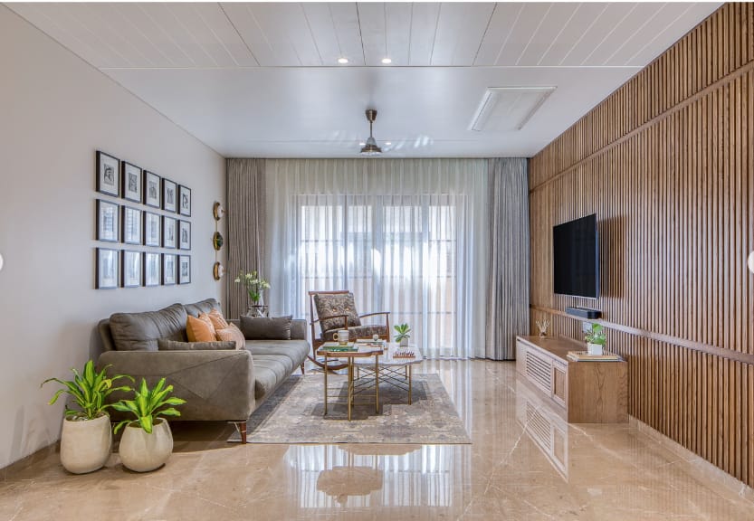 Modern living room interior design by Olangana in Bangalore, featuring a comfortable grey sofa, a striking vertical wooden slat wall with a mounted TV, and polished marble flooring, creating a warm and inviting space.
