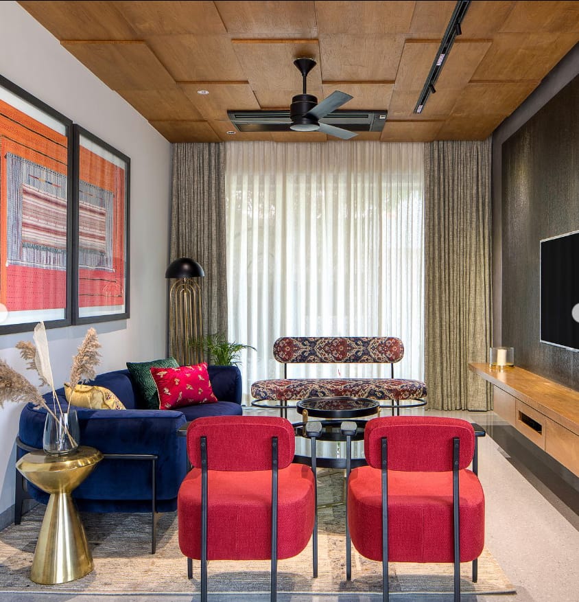 Modern living room interior design by Olangana in Bangalore, featuring a navy blue curved sofa, vibrant red accent chairs, a patterned bench, and a striking wood-paneled ceiling.