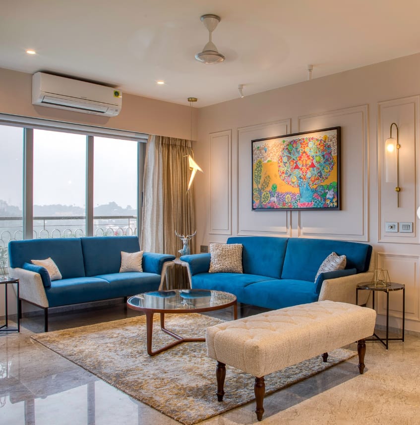 Elegant living room interior design by Olangana in Bangalore, featuring vibrant blue sofas, a stylish glass coffee table, a tufted beige bench, and a striking abstract art piece, complemented by modern lighting and a textured rug.