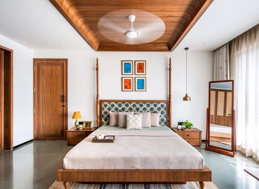 Elegant master bedroom interior design by Olangana Design in Bangalore, featuring a wooden four-poster bed with a patterned headboard, matching nightstands, and a unique wooden ceiling with a fan.