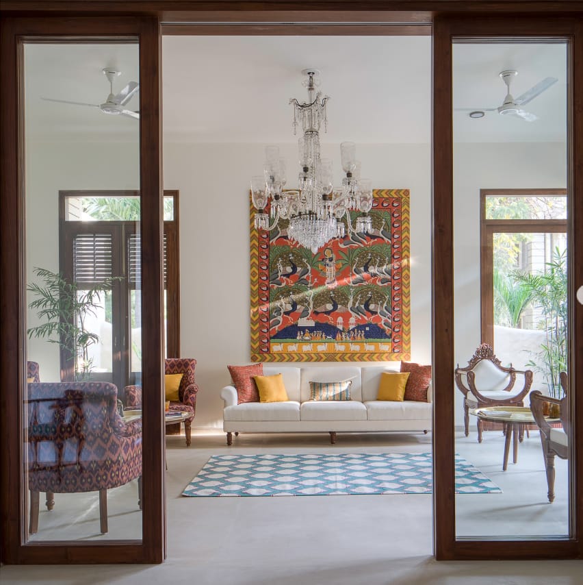 Elegant living room interior design by Olangana in Bangalore, featuring a stunning crystal chandelier, vibrant traditional tapestry, and a comfortable cream sofa with colorful cushions.