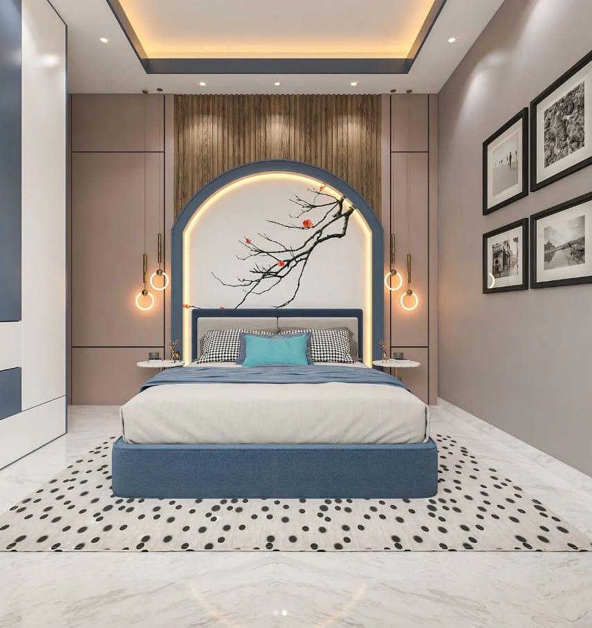 A stunning modern bedroom interior designed by Olangana Design in Bangalore, featuring a blue upholstered bed, an arched headboard with artistic tree branch motif, elegant pendant lights, and a patterned rug on marble flooring. This sophisticated interior showcases contemporary style with thoughtful design elements.
