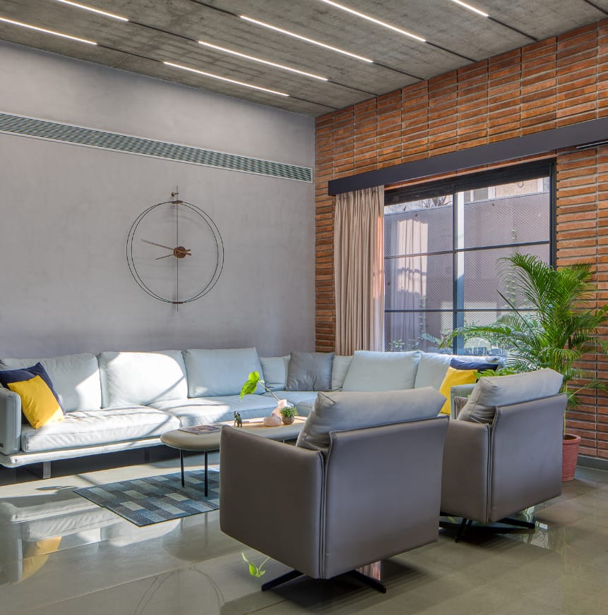 Modern industrial living room interior design by Olangana in Bangalore, featuring a light grey L-shaped sofa, exposed brick wall, concrete ceiling with linear lighting, and a minimalist wall clock.
