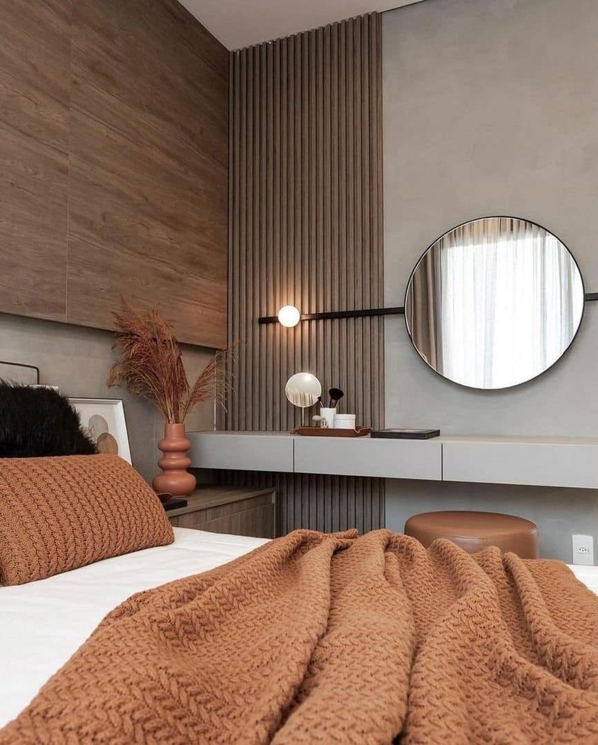 Modern bedroom interior design by Olangana Design in Bangalore featuring warm wood paneling, a sleek grey vanity with a large round mirror, and a cozy rust-colored knitted throw and pillow, creating a stylish and inviting space.