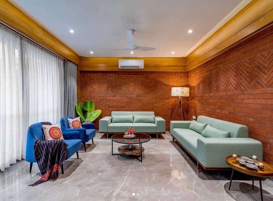 Modern living room interior design by Olangana in Bangalore, featuring a striking terracotta herringbone brick accent wall, comfortable mint green sofas, and vibrant royal blue armchairs, complemented by elegant lighting and marble flooring.