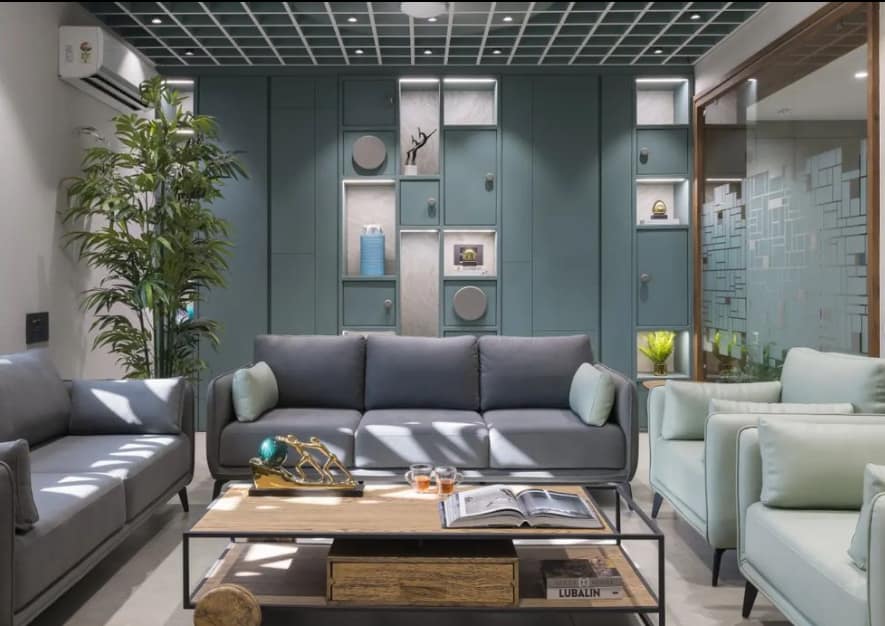 Modern living room interior design by Olangana in Bangalore, featuring a teal accent wall with integrated display shelves, comfortable grey and mint green sofas, and a stylish wooden coffee table.