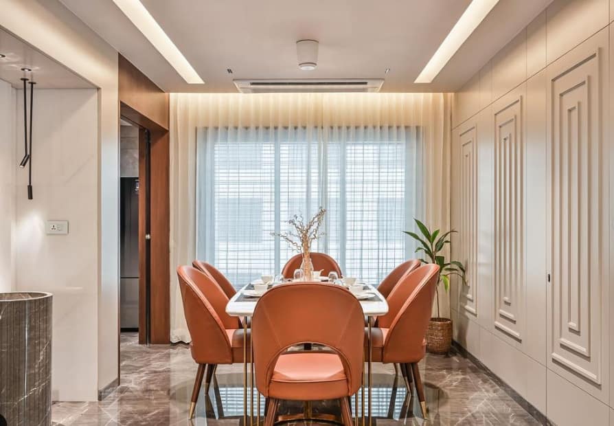 Elegant modern dining interior design by Olangana Design in Bangalore featuring burnt orange velvet chairs, a white marble dining table, and sophisticated lighting with decorative wall paneling.