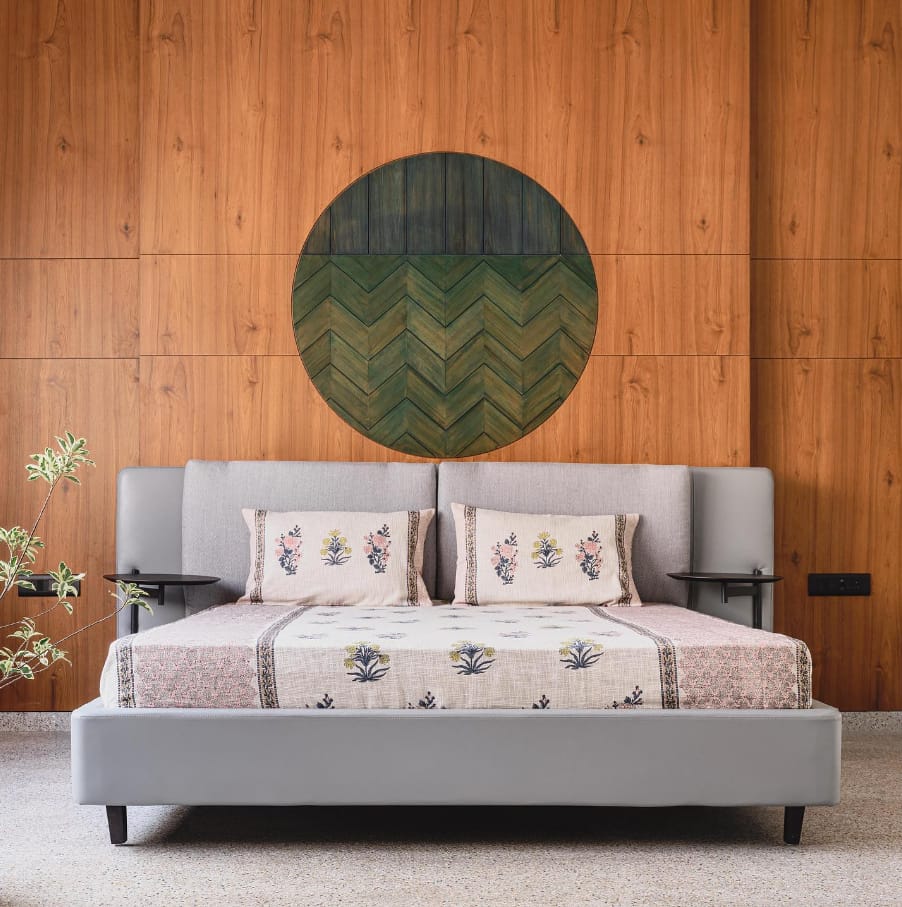 Elegant bedroom interior design by Olangana Design in Bangalore, showcasing a grey upholstered bed with floral bedding, floating bedside tables, and a distinctive circular green wood art installation against a paneled wall, reflecting premium interior aesthetics.