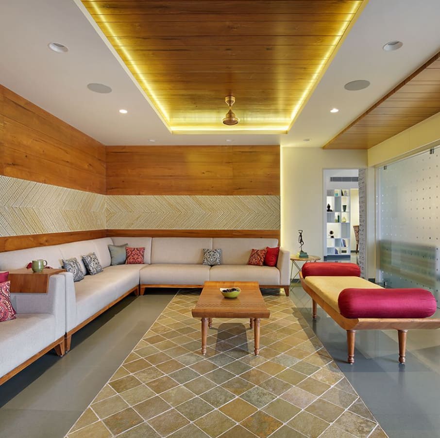 Modern living room interior design by Olangana in Bangalore, featuring a spacious L-shaped sofa, warm wooden ceiling with ambient lighting, and a distinctive patterned rug.