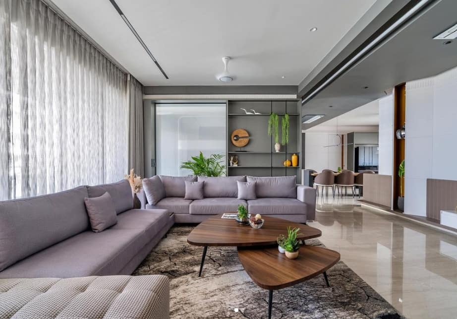 Modern grey living room interior design by Olangana in Bangalore, featuring a comfortable L-shaped sofa, unique triangular coffee tables, and stylish open shelving with plants.