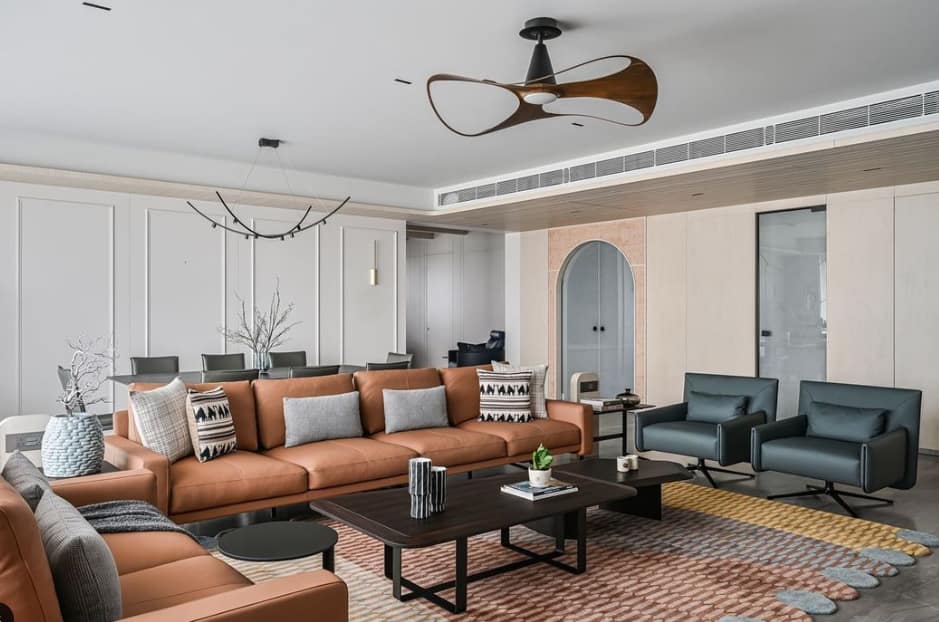 Modern open-plan living and dining room interior design by Olangana in Bangalore, featuring a tan leather sectional sofa, dark grey armchairs, a unique ceiling fan, and a vibrant geometric rug.