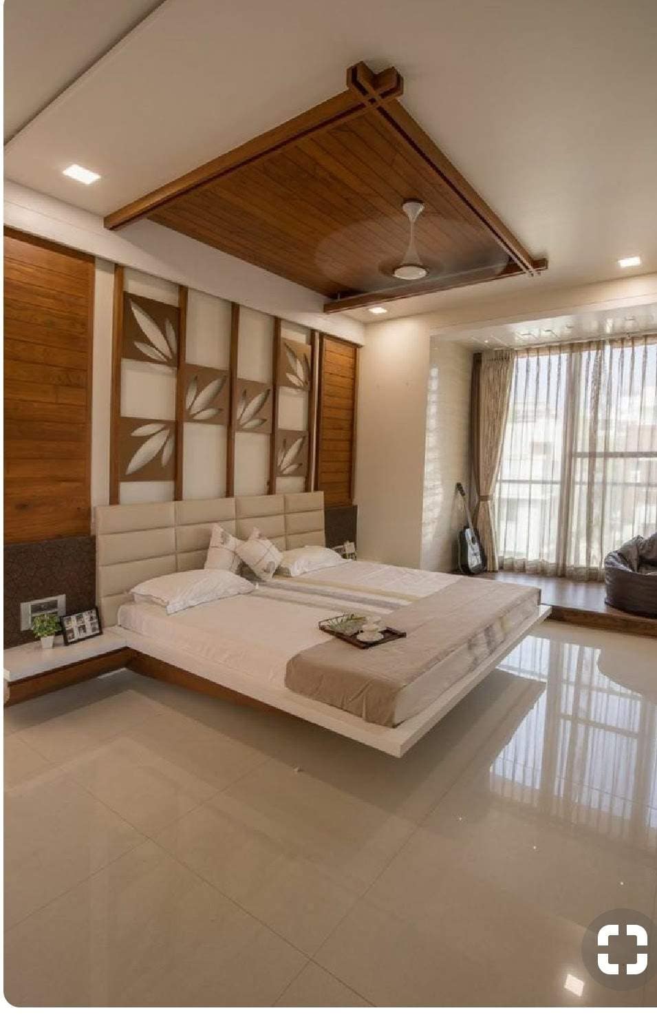 Modern bedroom interior featuring a large bed with a cushioned headboard, wooden wall paneling with leaf patterns, a wooden false ceiling, and elegant lighting, designed by Olangana Design in Bangalore.