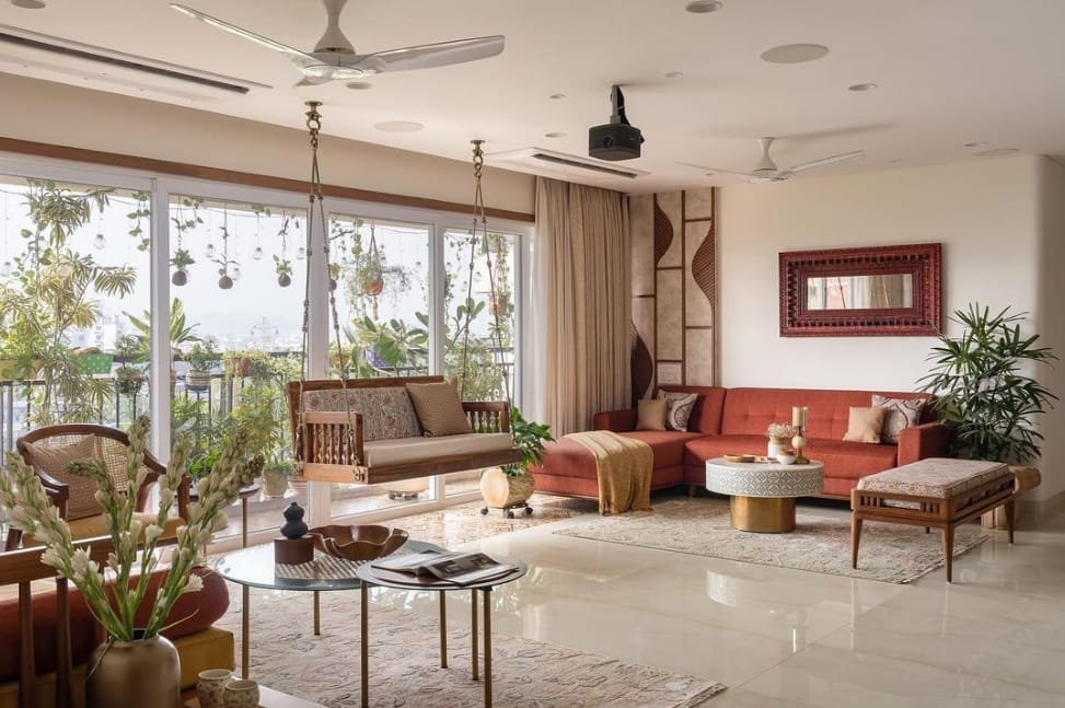 Elegant living room interior design by Olangana in Bangalore, featuring a warm terracotta sectional sofa, a traditional wooden swing, lush balcony greenery, and a carved mirror, creating a serene and inviting space.