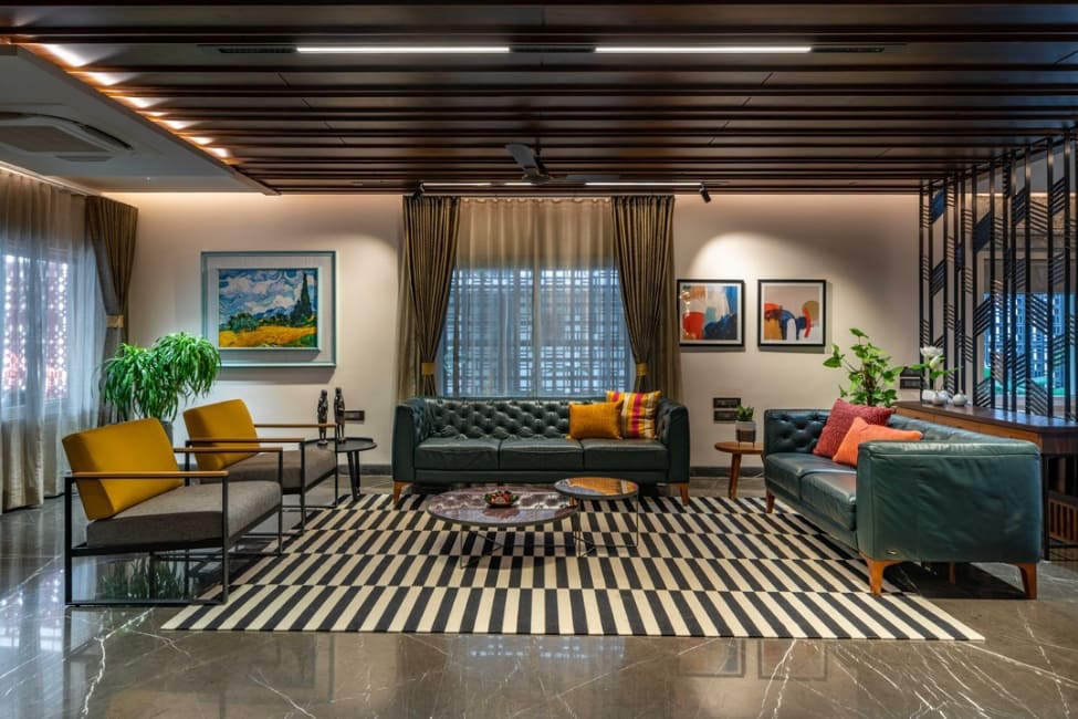 Elegant living room interior design by Olangana in Bangalore, featuring dark green tufted sofas, yellow accent chairs, a striking black and white striped rug, and warm ambient lighting.